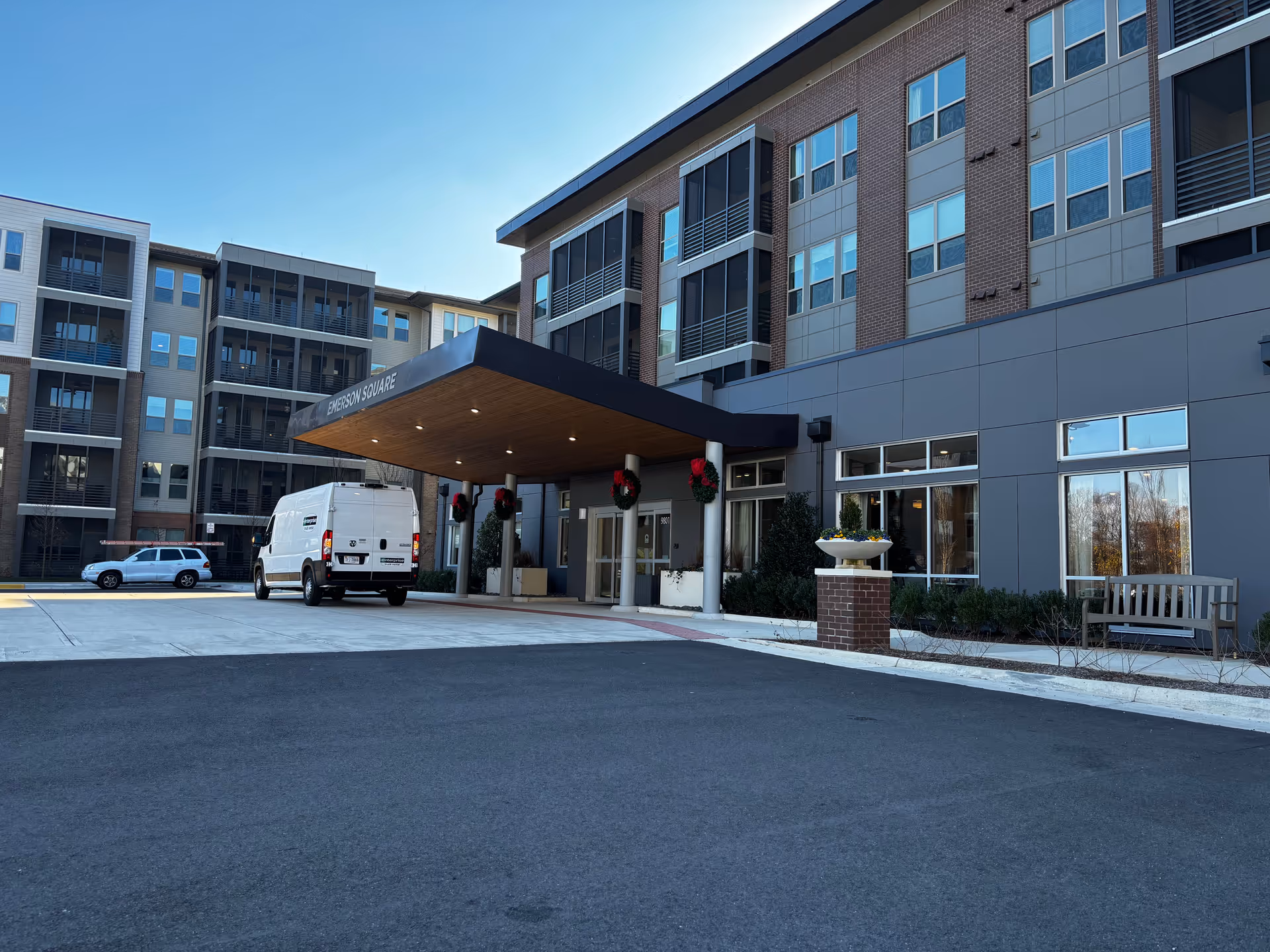 Exterior view of a modern senior living facility named Emerson Square with a covered entrance. There are two vehicles parked near the entrance, a white van and a white SUV. The building has multiple floors with large windows and balconies. There are holiday wreaths hanging on the pillars supporting the entrance canopy, and a bench and planter are visible near the entrance.