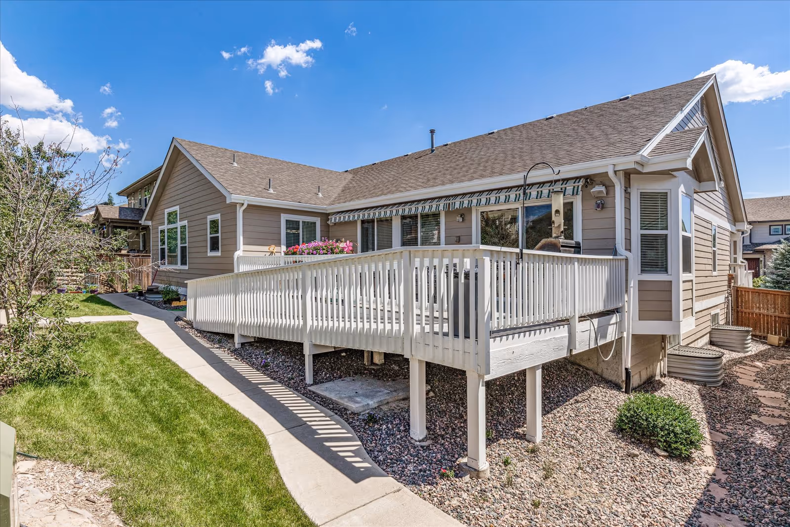 Exterior view of a single-story residential building with beige siding and a brown shingled roof. The building features a white wooden ramp leading to a patio area with a striped awning. The surrounding yard has green grass, a concrete walkway, and some landscaping with rocks and bushes under a blue sky with a few clouds.
