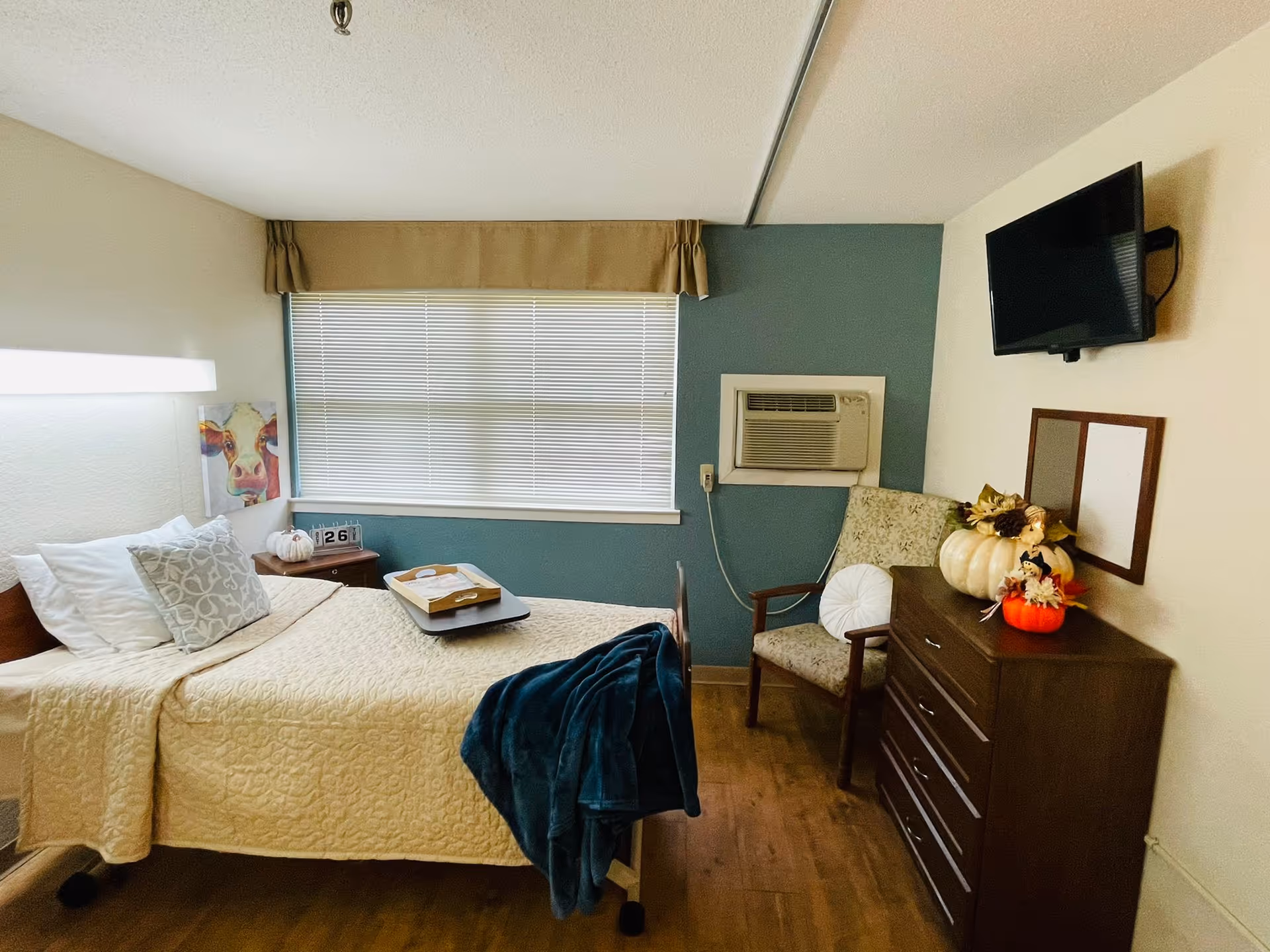A cozy bedroom in a senior living facility with a single bed covered in a beige quilt and pillows. A tray with a book and glasses rests on the bed. There is a window with closed blinds and a beige valance above it. Next to the window is a wall-mounted air conditioning unit. A wooden dresser with autumn-themed decorations, including pumpkins and flowers, is against the wall. A patterned armchair with a round white pillow is placed beside the dresser. A flat-screen TV is mounted on the wall above the dresser. The floor is wooden, and the room has a calm and welcoming atmosphere.