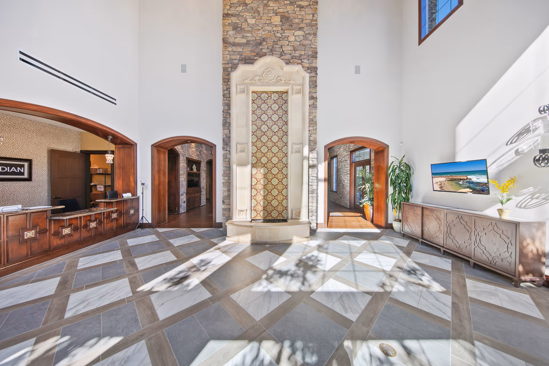 Bright two-story lobby with a central decorative tiled fountain, a wooden reception desk on the left, and a console with a TV on the right.