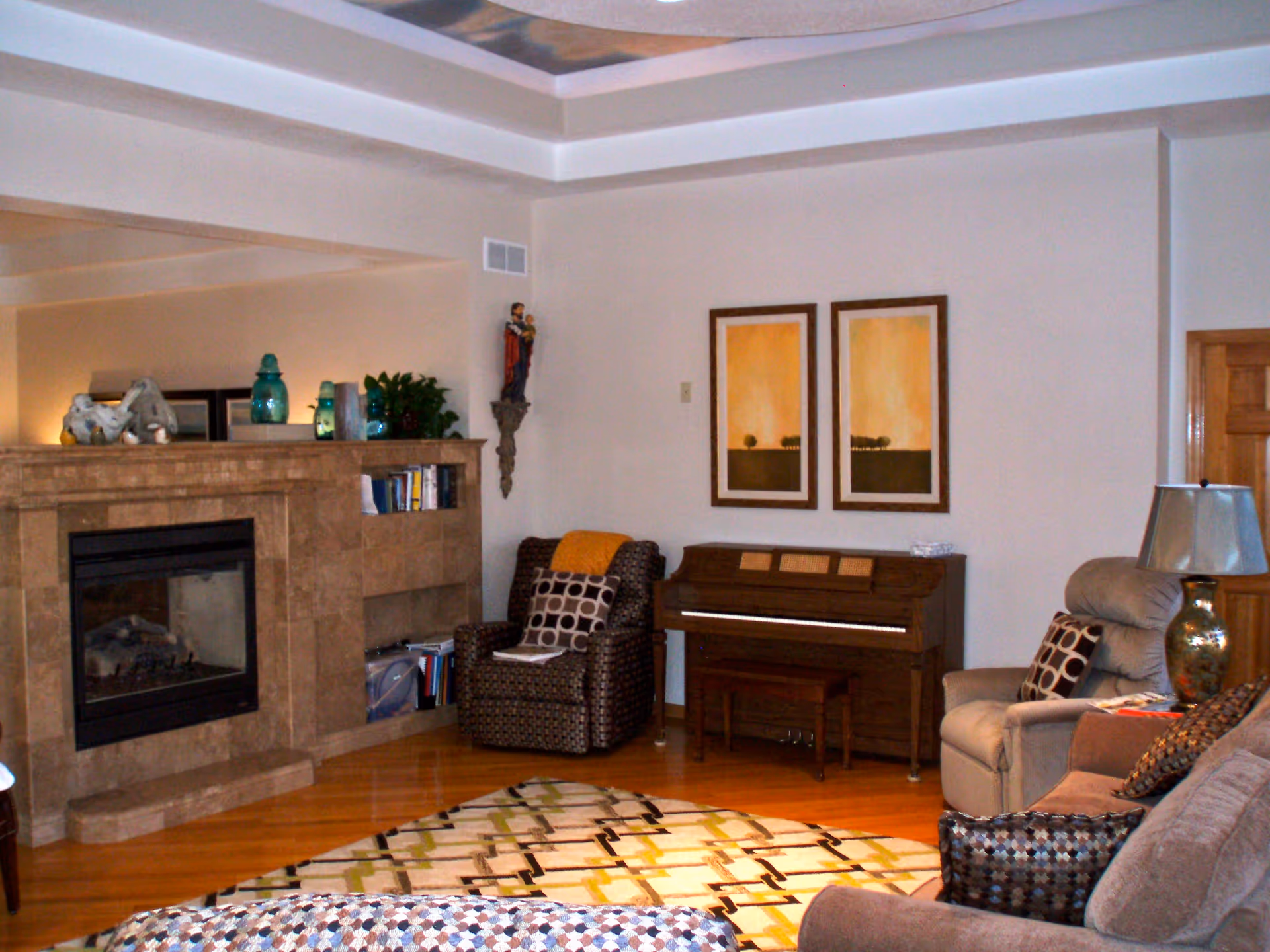 Cozy living room with a tiled fireplace, an upright piano against the wall, patterned armchairs and sofas, and a decorative rug on hardwood floors.