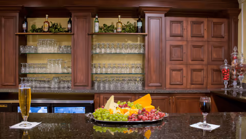 A bar area with wooden cabinets and shelves filled with glassware and bottles. On the counter, there is a large platter with assorted cheeses, grapes, and fruit. There are two drinks on the counter, one appears to be a glass of beer and the other a glass of red wine.