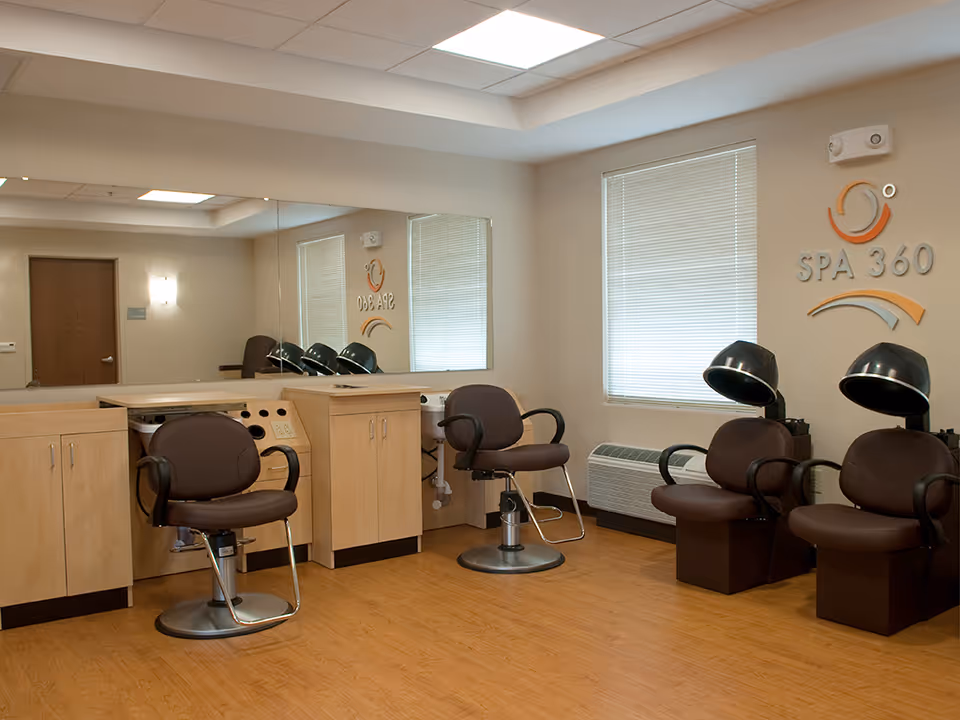 Interior view of a spa or salon area in BridgePort Healthcare Center with three brown salon chairs, two under hair dryers, wooden cabinets, a large mirror on the wall, a window with blinds, and a wall sign that reads 'SPA 360'.