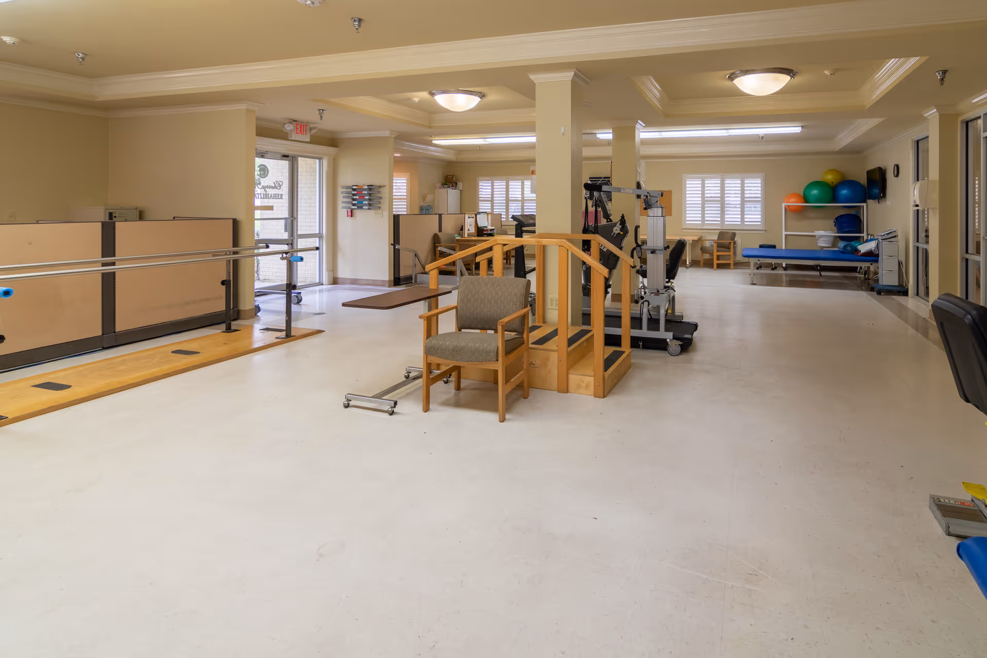 A spacious physical therapy room in a skilled nursing facility featuring parallel bars for walking exercises, a wooden staircase with handrails for step training, exercise equipment, therapy balls on a rack, and chairs arranged around the room. The room has beige walls, large windows with blinds, and bright overhead lighting.