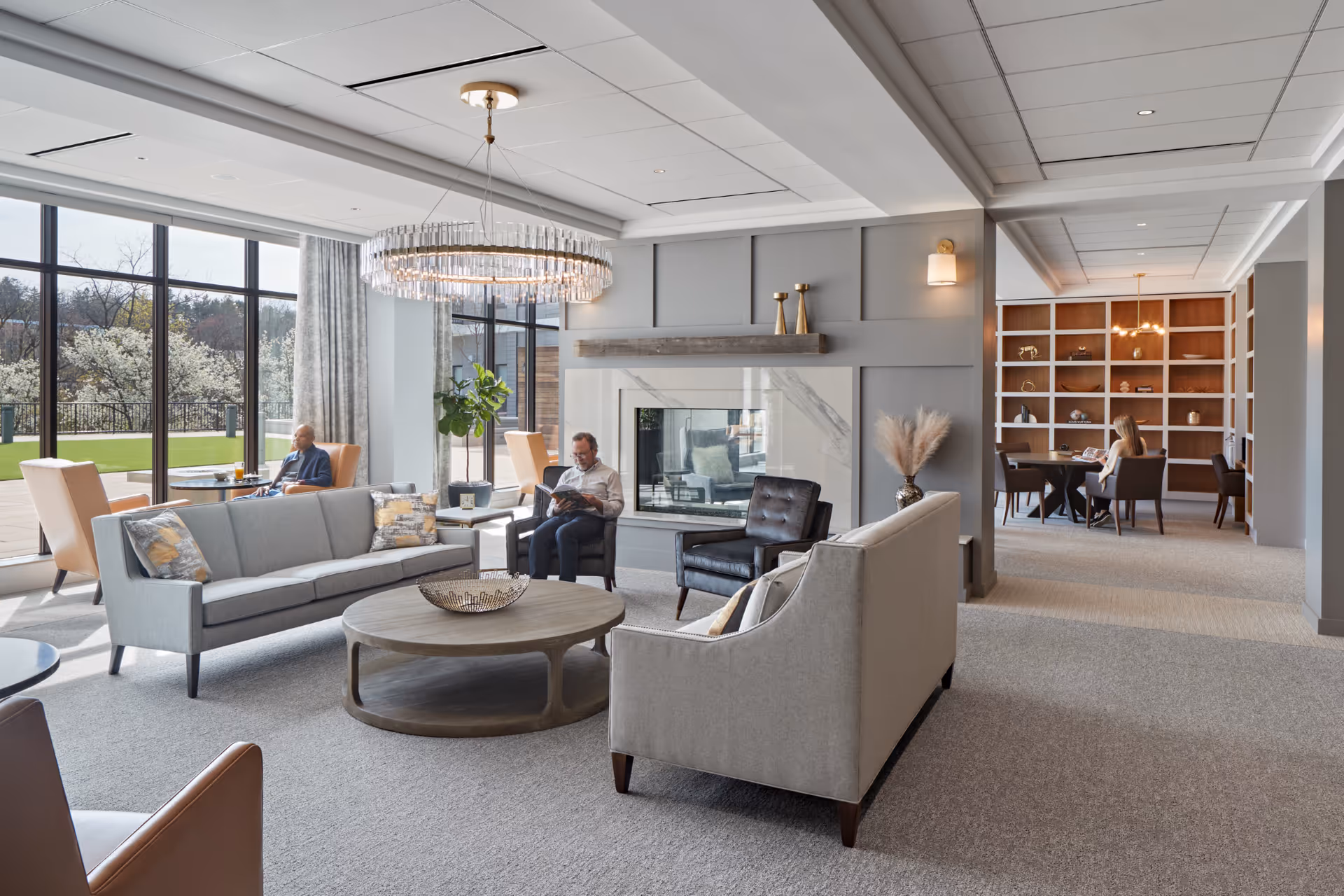 A spacious and modern living room area in a senior living facility with large windows showing an outdoor garden. The room features two gray sofas, two black armchairs, a round wooden coffee table, and a contemporary chandelier. There is a fireplace with decorative items on the mantel and a built-in bookshelf in the background. Two people are seated, one reading and the other looking outside, while a third person is seated at a table in the background.