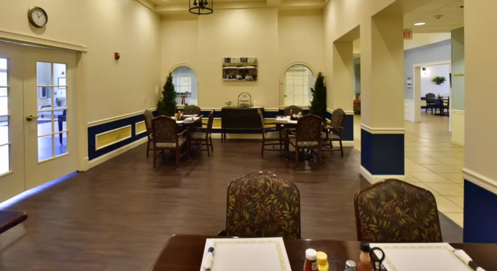 Communal dining room with multiple tables and chairs, placemats and condiments on a foreground table, wood flooring and double doors to the left.