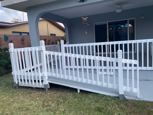 White ramp with railing leading up to a covered patio and sliding glass doors on a single-story house with a small lawn.