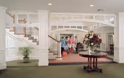 Spacious and elegant interior lobby area of a senior living facility with white paneled walls, a staircase with wooden handrails, a round table with a large floral arrangement in the center, and three people standing near the back entrance.