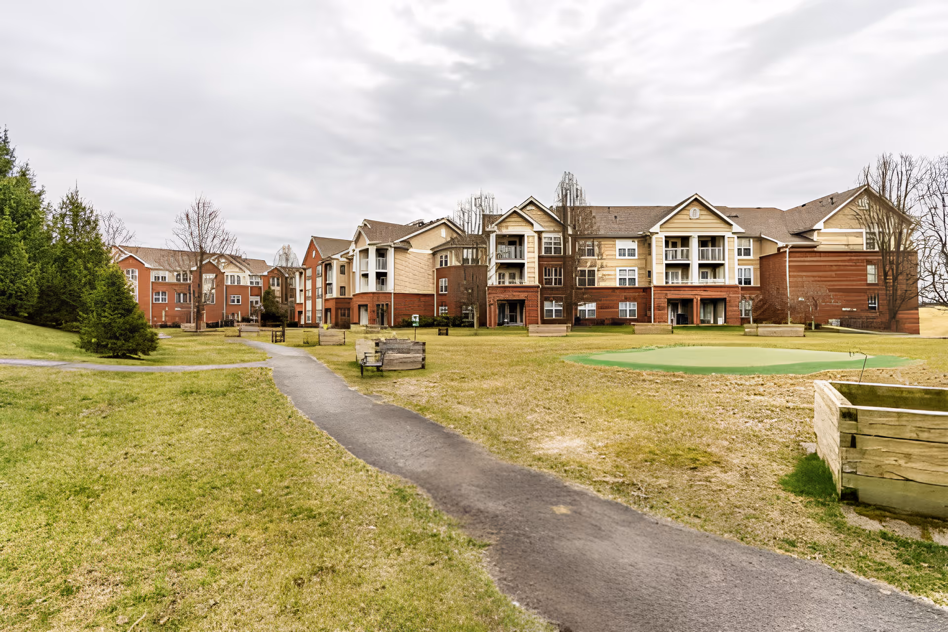 Exterior view of a multi-story senior living facility building with a paved walkway leading through a grassy area with benches and trees under a cloudy sky.