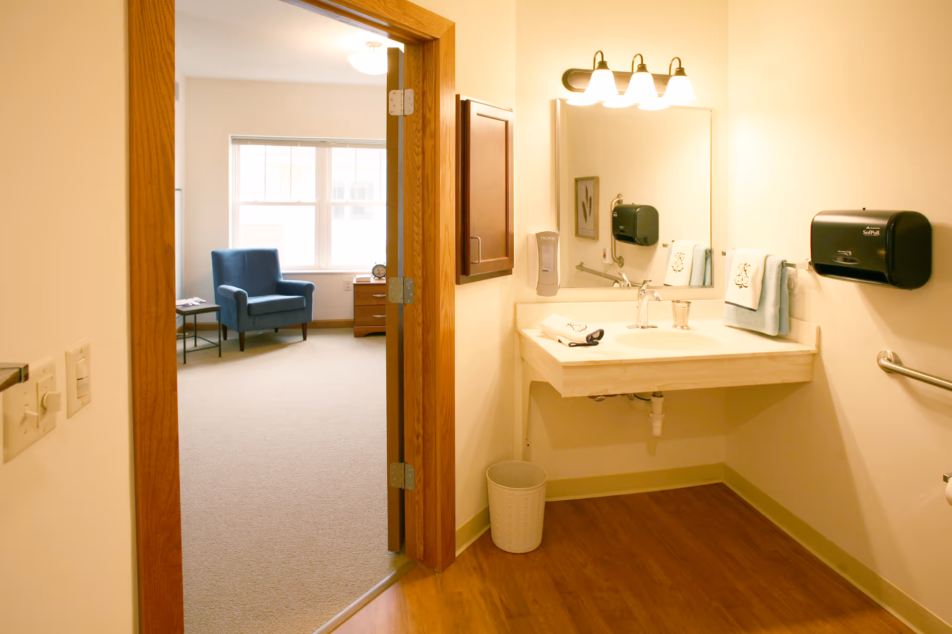 View of a bathroom with a sink, mirror, hand soap dispenser, paper towel dispenser, and handrails. The bathroom has wooden flooring and beige walls. Through an open door, a room with a blue armchair, side table, and window with blinds is visible.