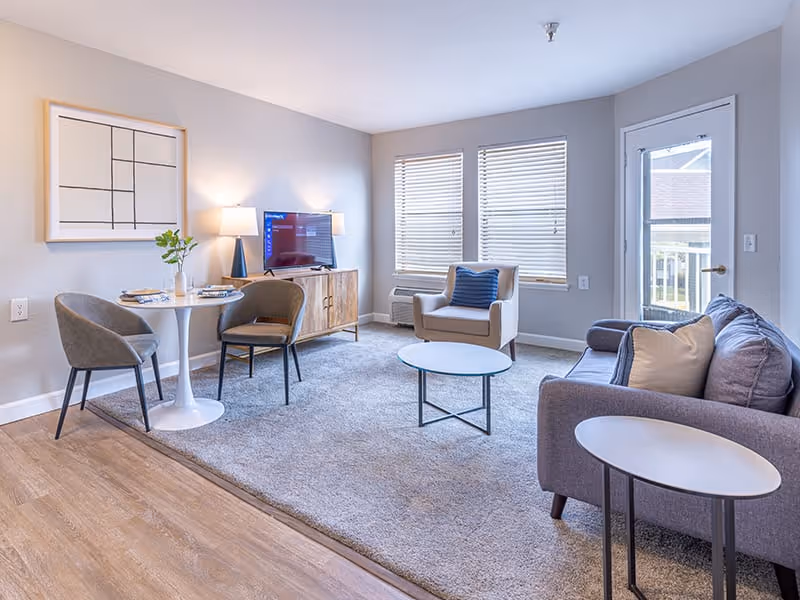 A bright living room with a gray sofa, a beige armchair with a blue pillow, a round coffee table, and a small round dining table with two gray chairs. A wooden TV stand holds a flat-screen TV and a lamp. Two windows with blinds and a glass door leading outside are visible.
