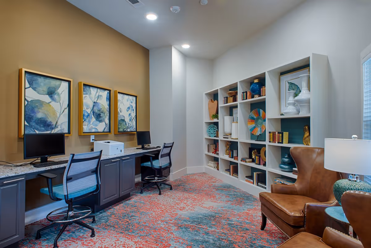 A cozy interior room featuring a workspace with two computer desks and chairs along a wall with three framed abstract blue and green paintings. Opposite the desks is a white shelving unit filled with decorative items and books. In the foreground, there is a brown leather armchair next to a small round table with a green lamp. The room has a colorful patterned carpet and recessed ceiling lights.