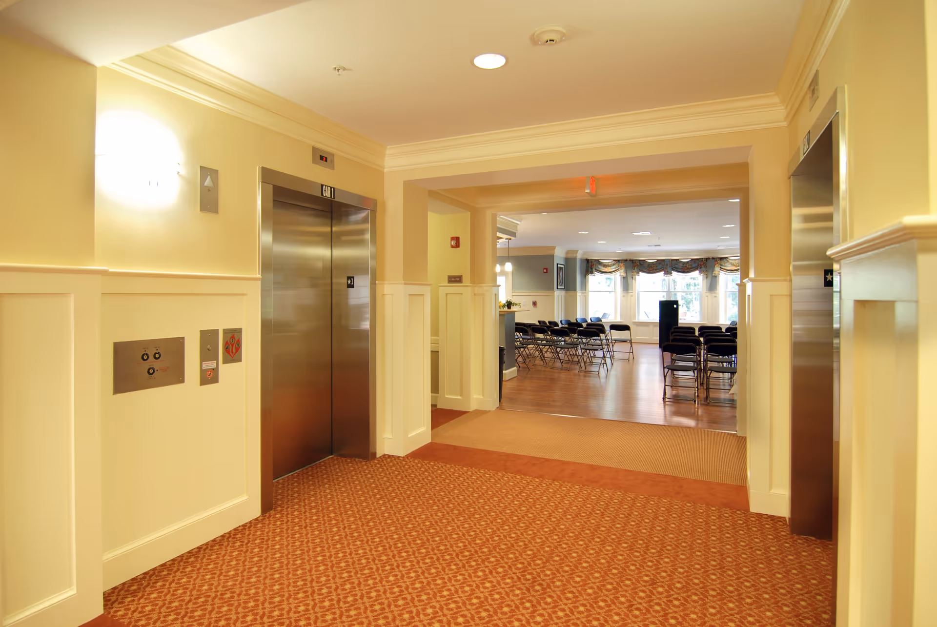 Interior view of a senior living facility hallway with two stainless steel elevator doors on either side. The hallway has beige walls with white wainscoting and a patterned red carpet. Beyond the hallway is a room with wooden flooring and rows of black folding chairs arranged facing a speaker podium near large windows with floral valances.