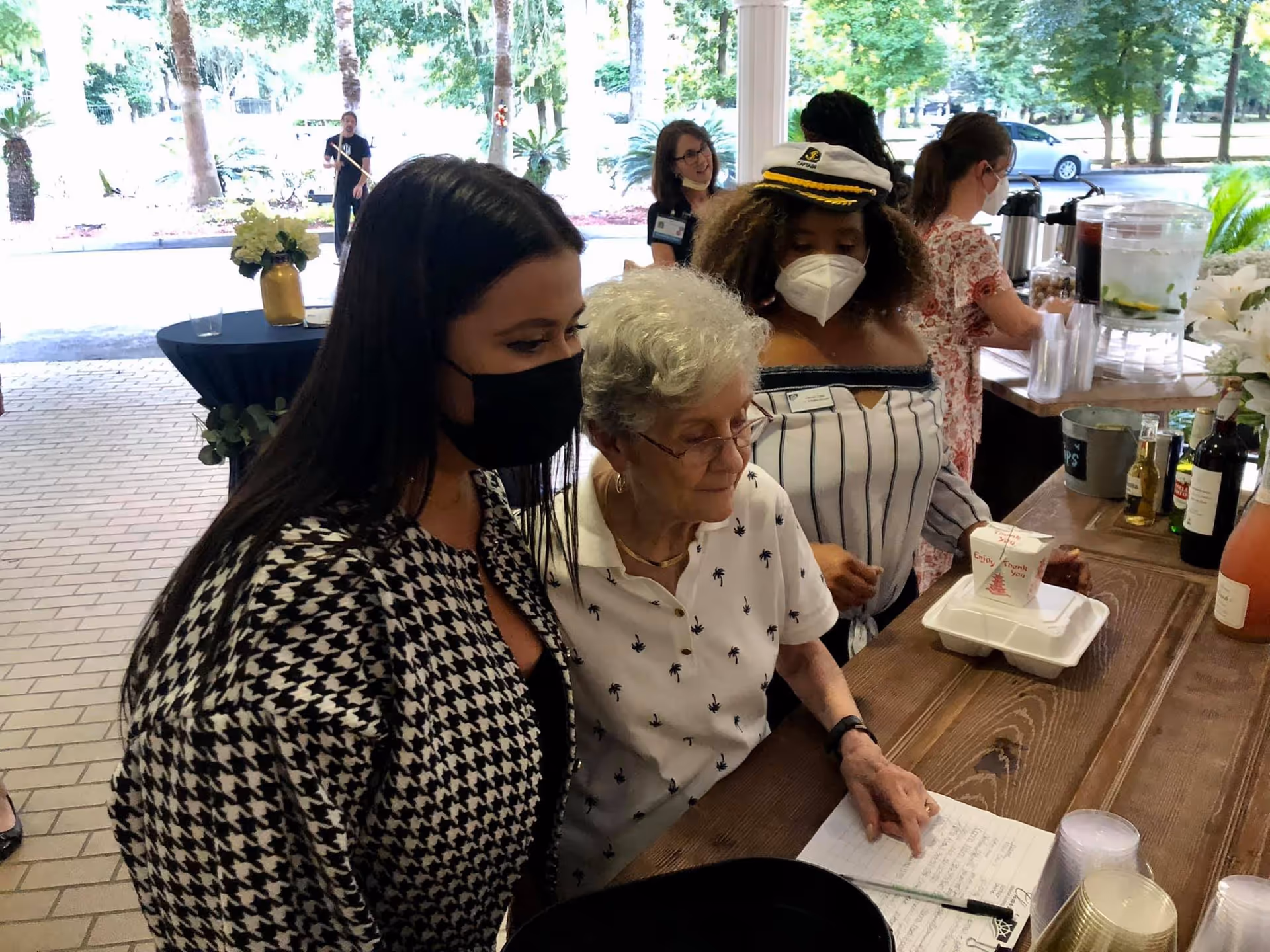 A group of people gathered around a wooden counter outdoors. An elderly woman with white hair and glasses is pointing at a notebook on the counter. Two women wearing face masks stand beside her, one in a black and white houndstooth jacket and the other in a striped off-shoulder top with a captain's hat. In the background, a woman is serving drinks from a beverage station with cups and bottles visible. Trees and a parked car are seen outside.