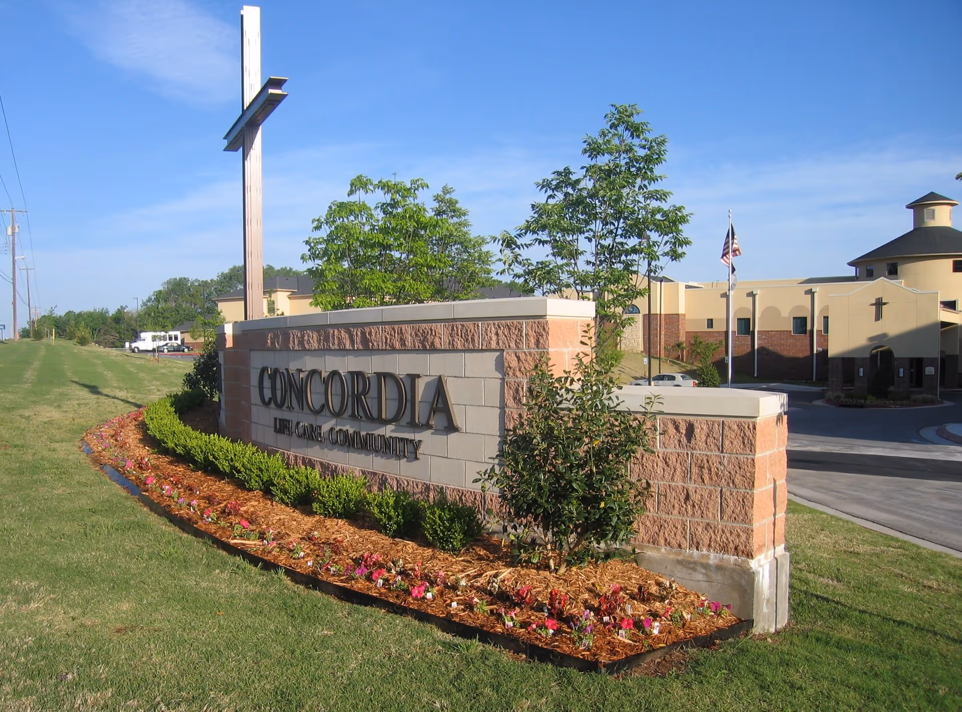 Outdoor view of the entrance sign for Concordia Life Care Community, featuring a large cross mounted on a brick and stone wall with landscaping and flowers in front. The facility building and an American flag are visible in the background under a clear blue sky.
