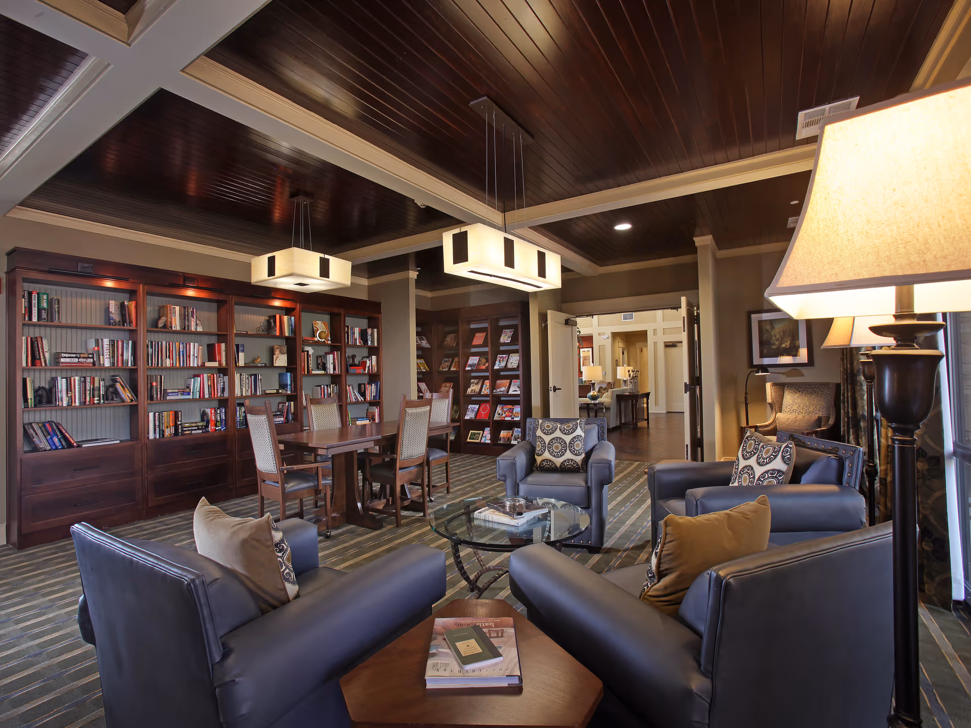 A cozy and well-lit common area with dark leather armchairs arranged around a glass coffee table. Behind the seating area, there is a wooden table with chairs and bookshelves filled with books and magazines. The ceiling is dark wood with modern rectangular pendant lights hanging down. A floor lamp is visible on the right side, and the room has a warm and inviting atmosphere.