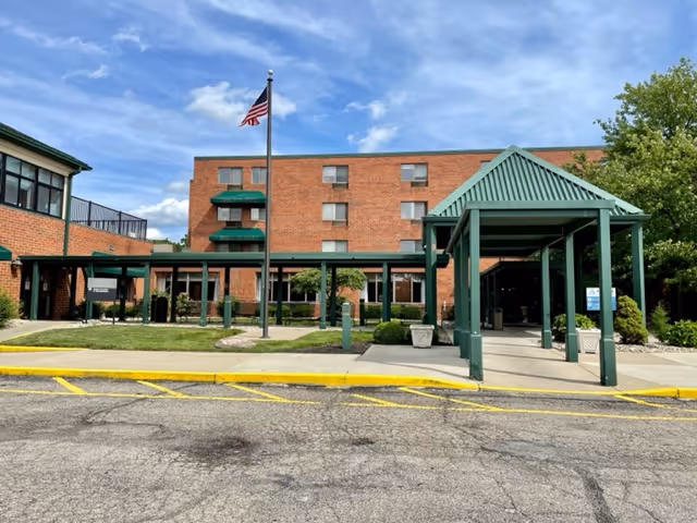 Front entrance of a brick senior living building with a green covered portico, flagpole, and landscaped courtyard.