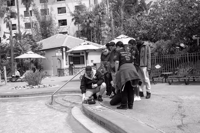 A group of people gathered around the edge of a swimming pool outdoors, with one person crouching and pointing at something near the pool steps. There are lounge chairs, umbrellas, and a small building in the background surrounded by trees and plants.
