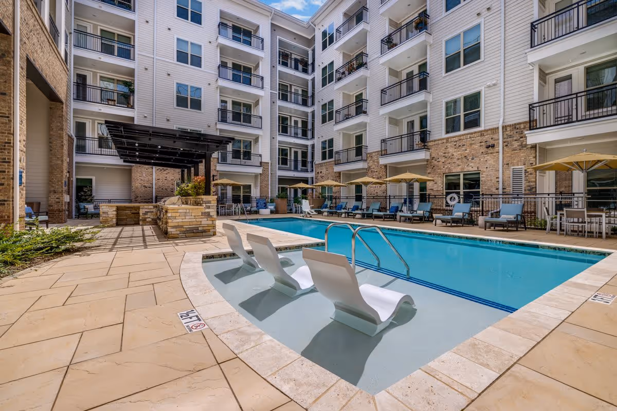 Outdoor swimming pool area in a multi-story residential or senior living facility with lounge chairs partially submerged in the shallow end of the pool, additional lounge chairs and umbrellas along the poolside, and a covered seating area with stone pillars. The building surrounding the pool has balconies and windows.