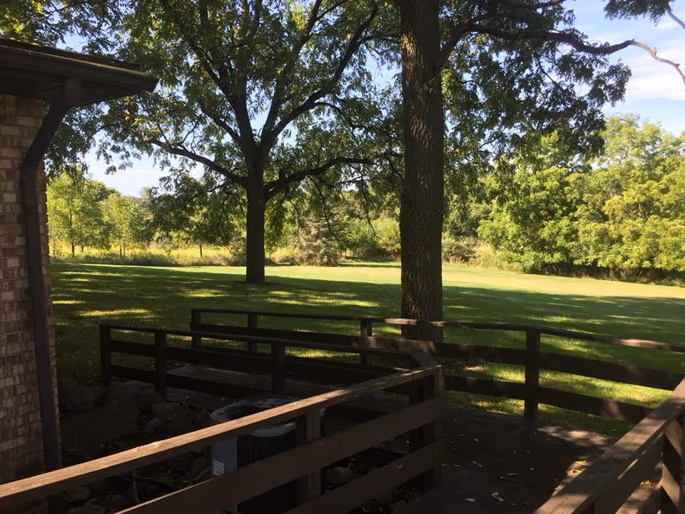 View of a green outdoor area with large trees and a grassy lawn, seen from a wooden deck attached to a brick building.