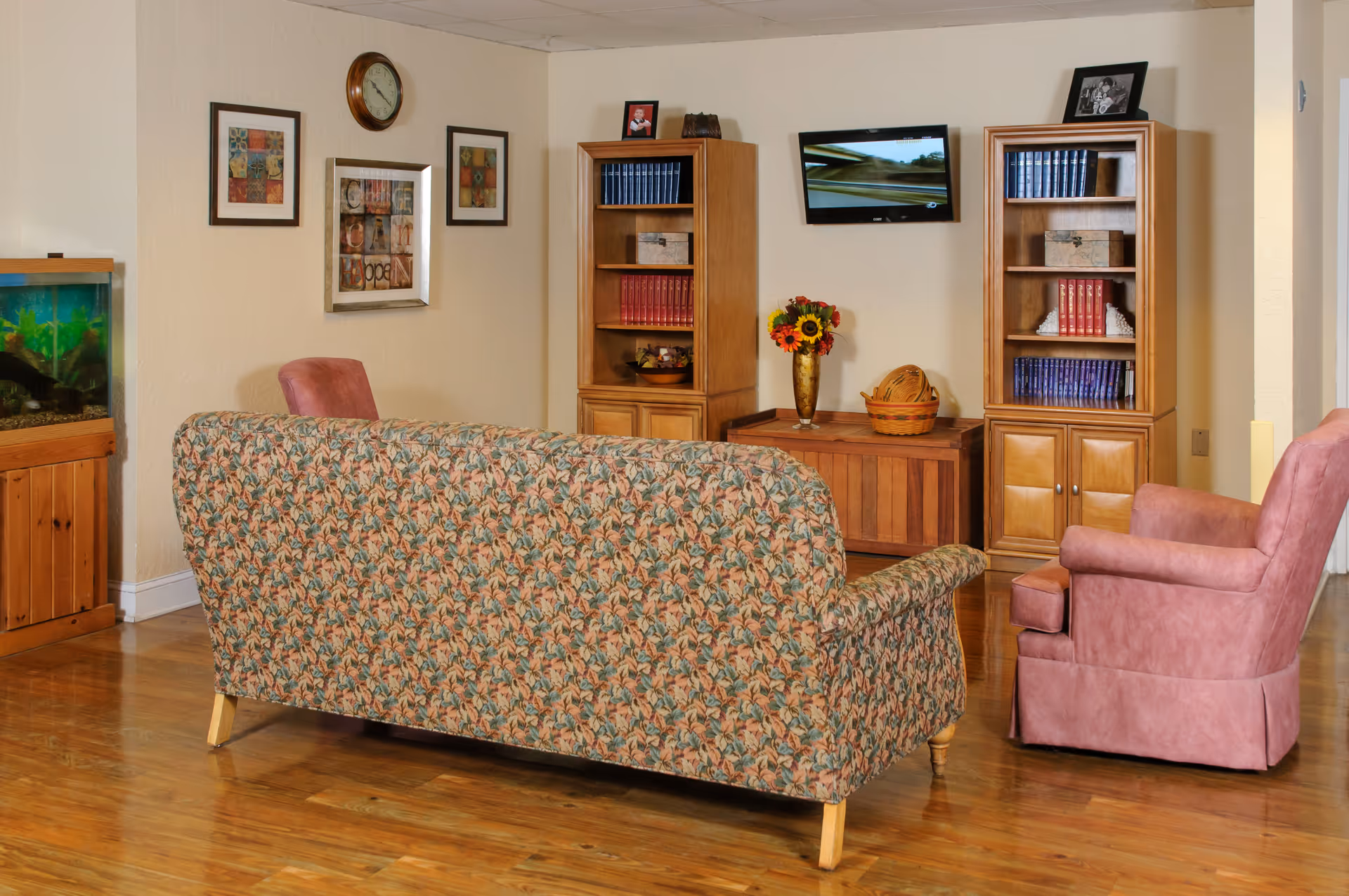 A cozy living room area with a floral patterned sofa and two pink armchairs facing a wall-mounted TV. The room has wooden flooring, two wooden bookshelves filled with books and decorative items, a wooden cabinet with a vase of flowers and a basket on top, framed pictures and a clock on the wall, and a fish tank on the left side.