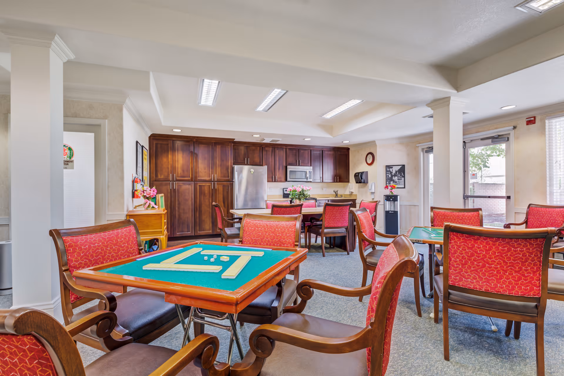 A bright and spacious common area in a senior living facility featuring several wooden tables with green felt tops set up for games, surrounded by red cushioned chairs. In the background, there is a kitchen area with dark wooden cabinets, a stainless steel refrigerator, microwave, and a sink. The room has large windows and glass doors letting in natural light, with a water dispenser and wall decorations visible.