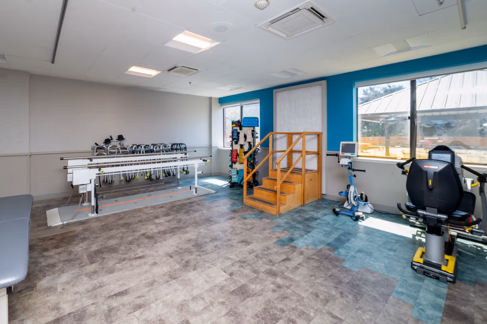 A rehabilitation therapy room with parallel bars for walking exercises, a set of wooden stairs for physical therapy, exercise equipment including a recumbent bike, and a rack holding multiple walkers. The room has large windows letting in natural light and a blue accent wall.