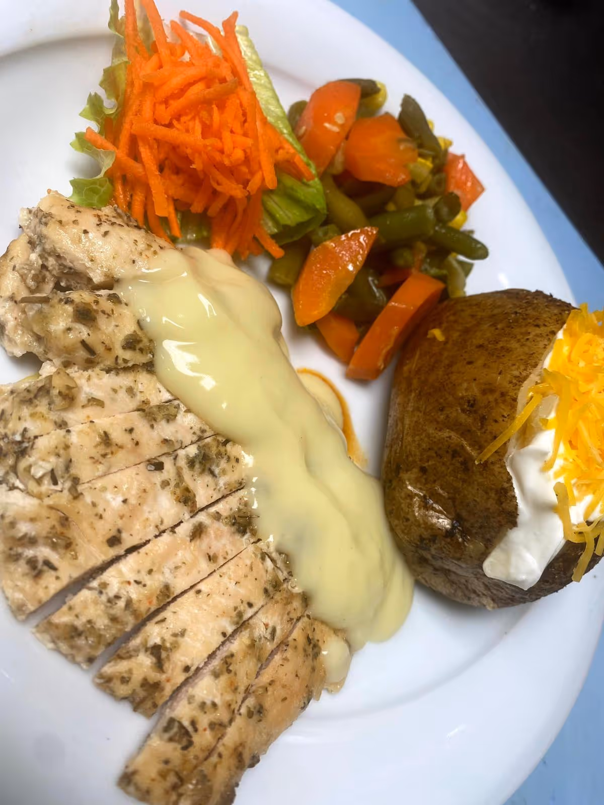 A plate of food featuring sliced herb-seasoned chicken breast topped with a creamy sauce, a baked potato with sour cream and shredded cheddar cheese, steamed mixed vegetables including carrots and green beans, and a small serving of shredded carrots on a lettuce leaf.