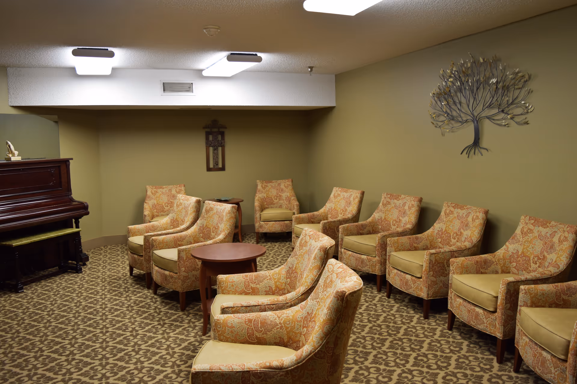 A cozy lounge with patterned armchairs arranged in rows, small tables, a piano, and wall decor on muted green walls.