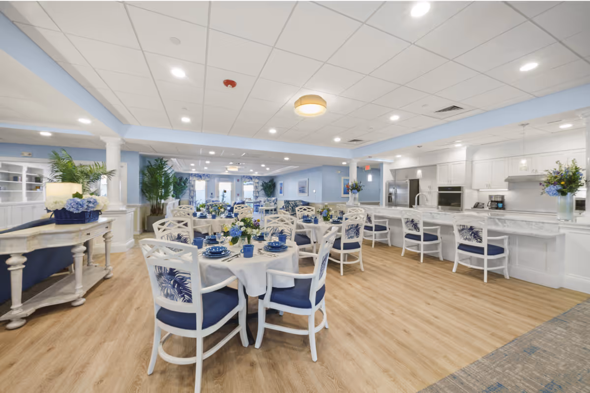 A bright and spacious dining room with several round tables set with blue and white dishware and floral centerpieces. The room features white chairs with blue cushions and leaf-patterned backs, light wood flooring, and a white kitchen area with a marble countertop and bar stools. The walls are painted light blue, and there are plants and decorative elements throughout the space.