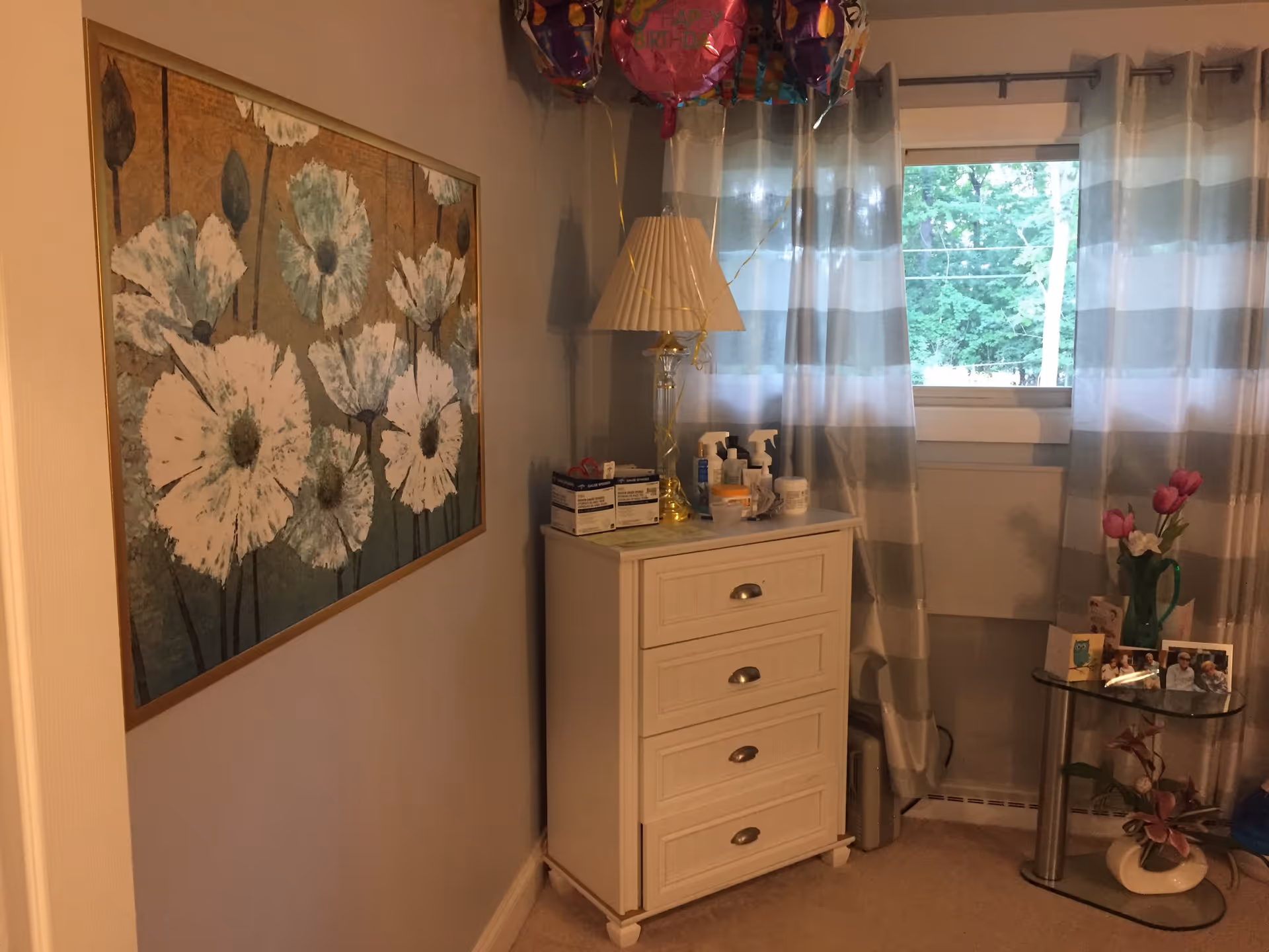 A cozy corner of a room featuring a white chest of drawers with various personal care items and a lamp on top. Next to it is a glass side table with a green pitcher holding pink flowers, framed photos, and a small plant underneath. A window with striped curtains lets in natural light, and a large floral painting hangs on the adjacent wall.