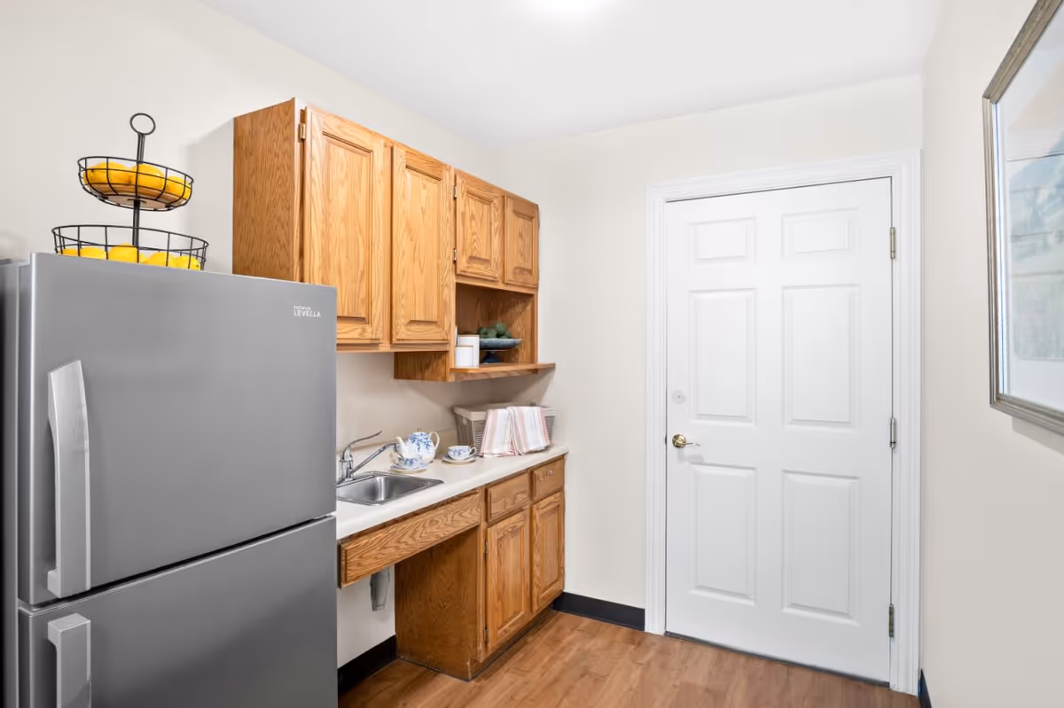 A small kitchen area with wooden cabinets, a stainless steel refrigerator, a sink, and a countertop with a tea set and towels. There is a white door and a framed picture on the wall.