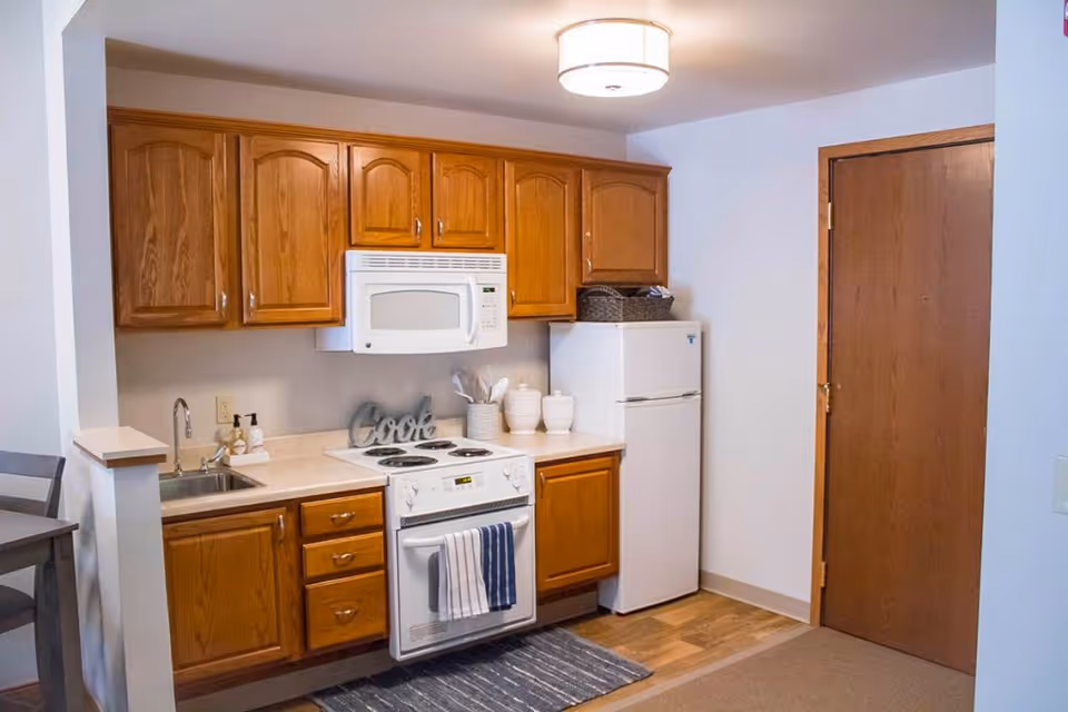 A small kitchen area with wooden cabinets, a white microwave above a white electric stove with a striped towel hanging on the oven door, a white refrigerator, a sink with soap dispensers, and a wooden door to the right. There is a small rug on the floor in front of the stove.