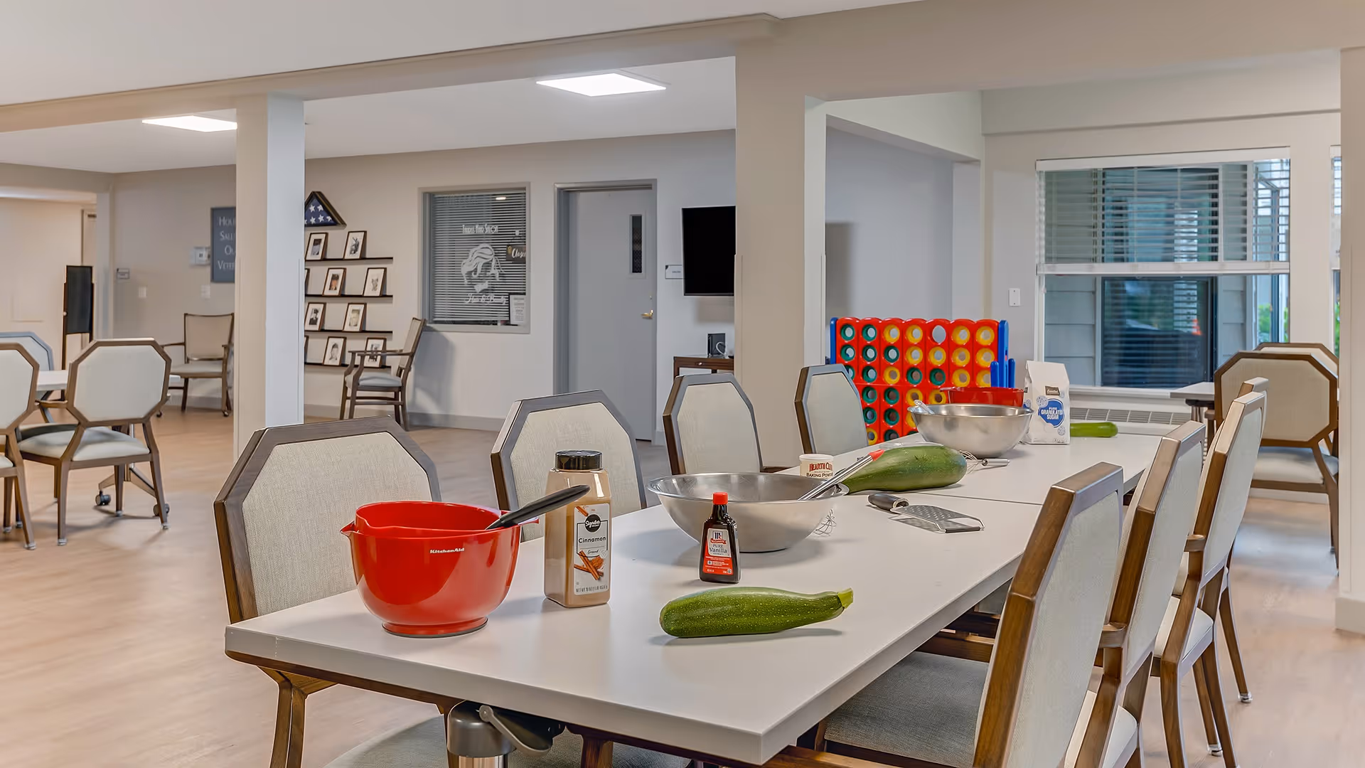 A bright and spacious common area with a long white table surrounded by chairs. On the table are mixing bowls, a red mixing bowl with a spoon, two zucchinis, a bottle of vanilla extract, a container of cinnamon, a grater, and a bag of flour. In the background, there are more chairs, a large Connect Four game, windows with blinds, and framed pictures on the walls.
