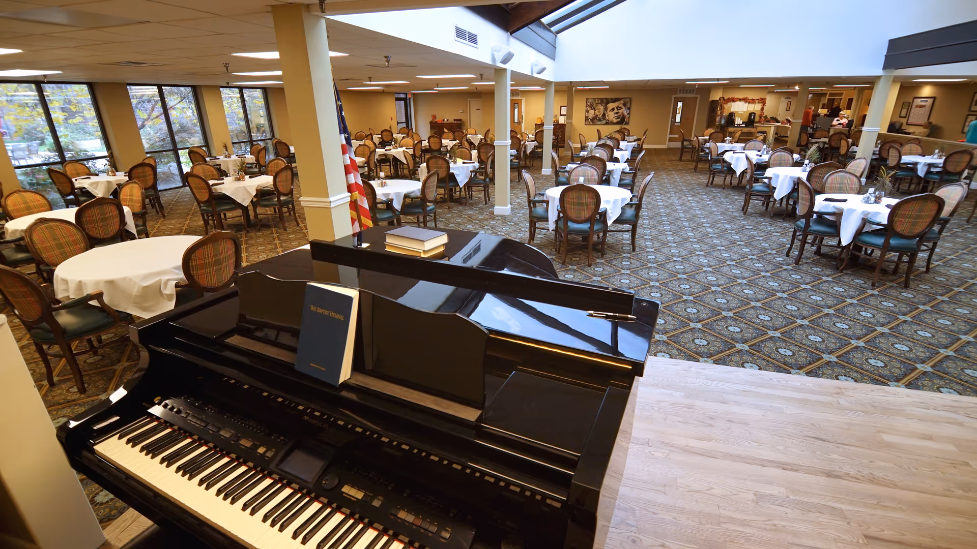 Interior view of a senior living community dining room with multiple round tables covered with white tablecloths and surrounded by wooden chairs with cushioned seats. A black grand piano with sheet music is in the foreground, and large windows on the left side let in natural light. The room has patterned carpet flooring and an American flag near the piano.