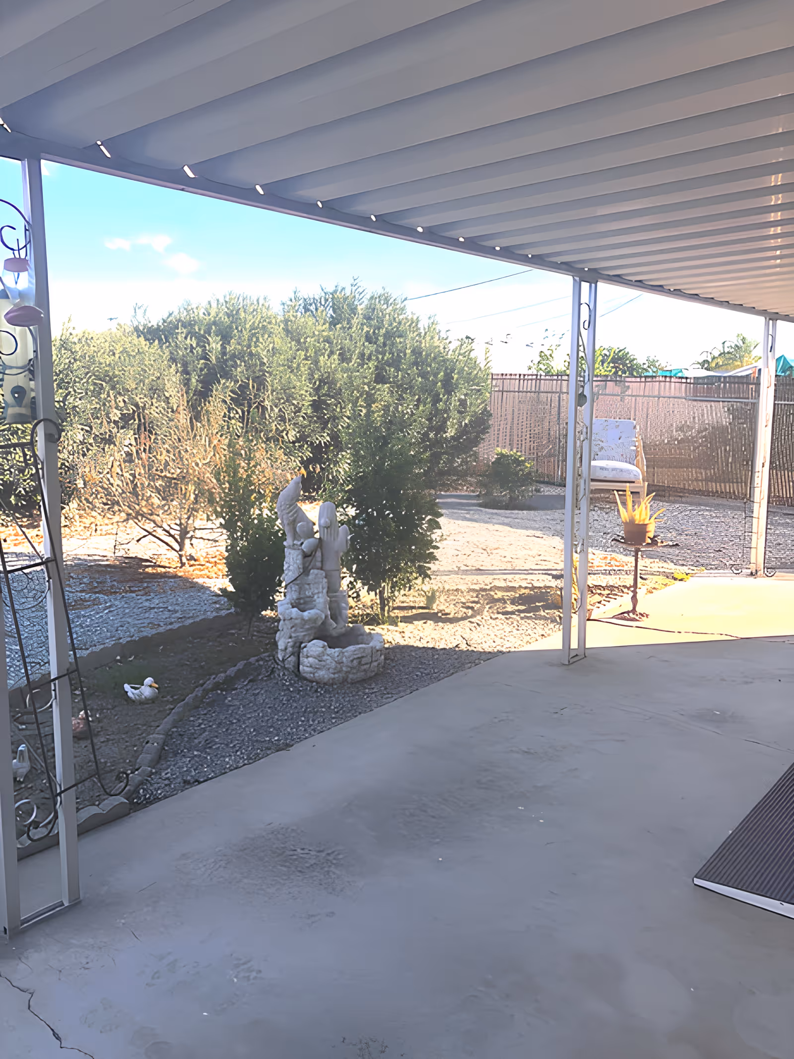 Covered patio area with a concrete floor overlooking a garden with bushes, a small stone fountain, and decorative items including a small duck figurine and a potted plant on a stand. There is a chair in the background near a fence.