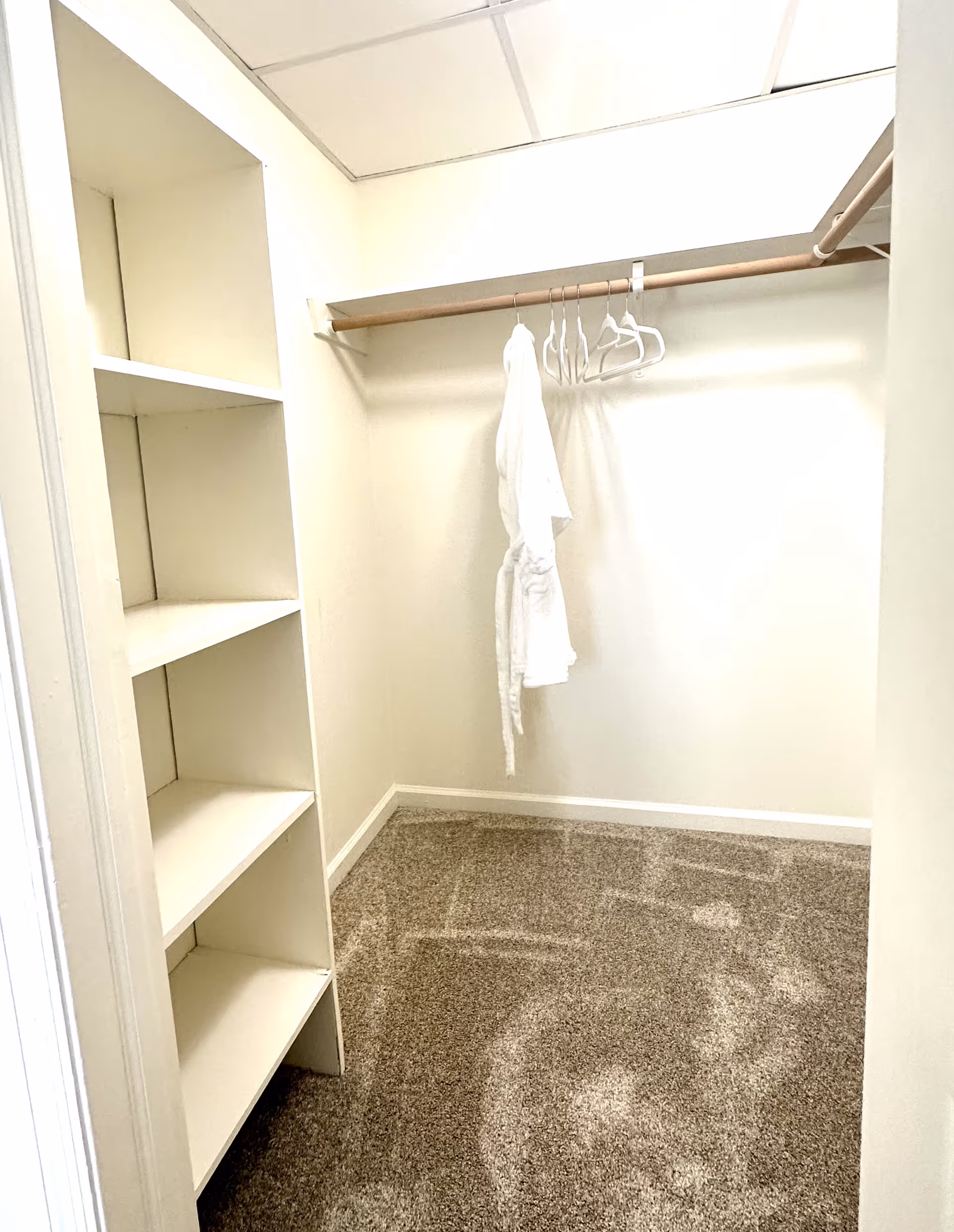 A small walk-in closet with beige walls and carpeted floor. There are built-in white shelves on the left side and a wooden hanging rod on the right with a few white hangers and a white robe hanging on it. The ceiling has white tiles with fluorescent lighting.