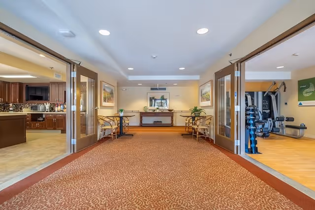 Interior view of a senior living facility hallway with patterned carpet, two sets of double glass doors on either side leading to a kitchen area on the left and a fitness room on the right. The hallway has chairs and small tables along the walls, framed artwork, and a console table with plants and a mirror at the far end.