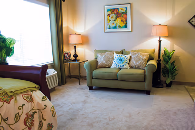 A bright bedroom with a green loveseat between two floor lamps, a bed in the foreground, and a colorful painting on the wall.