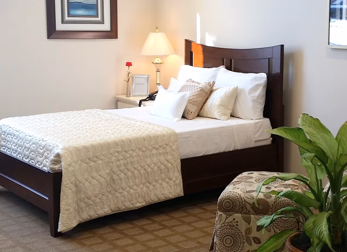 A furnished bedroom with a wooden bed dressed in white bedding and pillows, a nightstand with a lamp and framed art, and a potted plant.