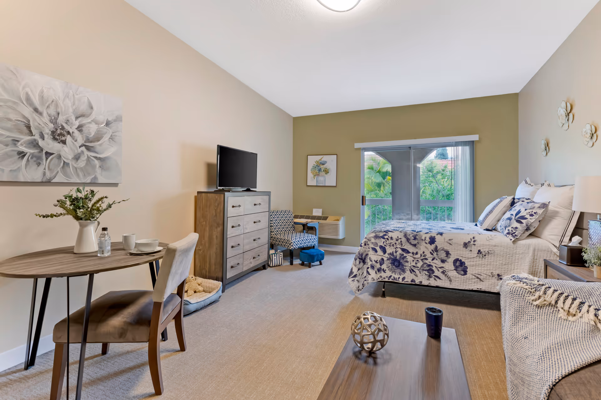Bright studio-style bedroom with a bed, dresser, small dining table and sliding glass door to a balcony.