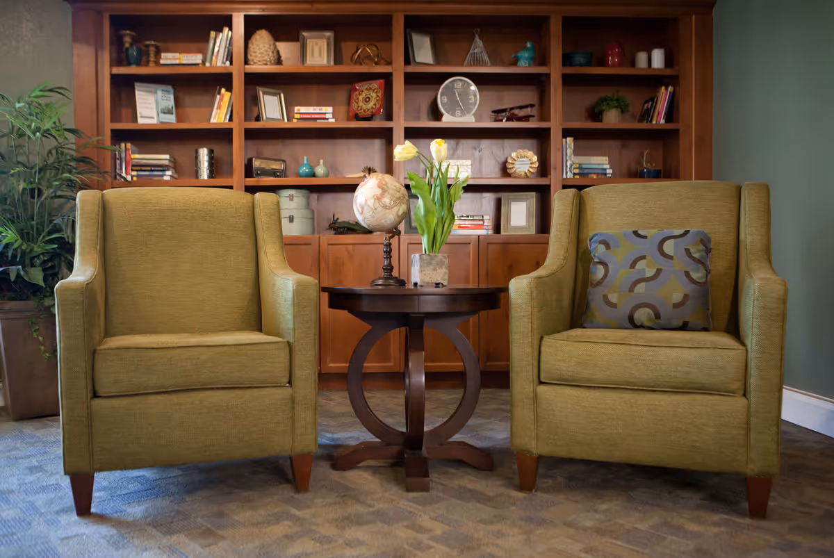 Two green upholstered armchairs with a round wooden side table holding a vase of flowers, set in front of a built-in bookshelf.