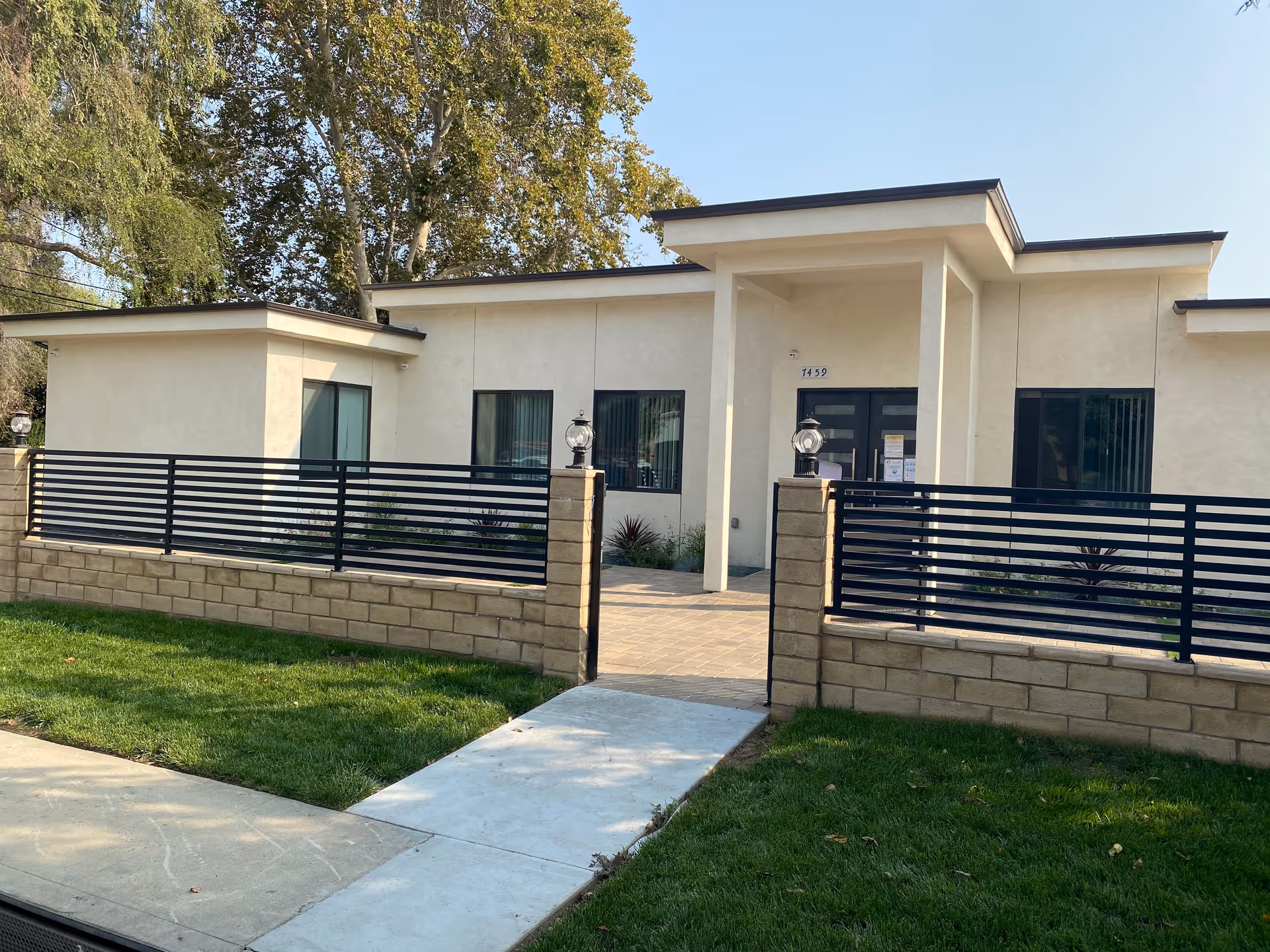 Front exterior view of a single-story building with a beige facade, black horizontal metal fence, and a paved walkway leading to the entrance. The entrance has a small covered porch with two columns and black double doors. There are several windows with vertical blinds and some greenery around the building.