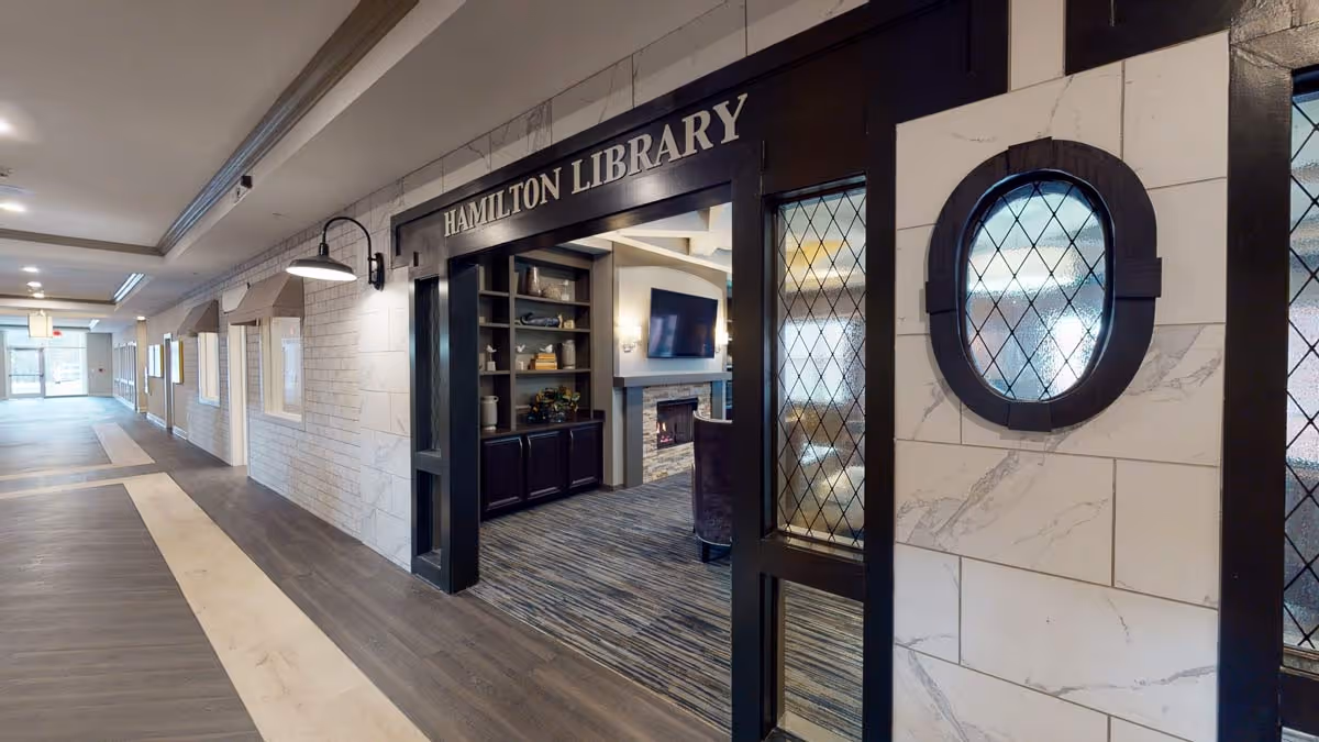 Interior hallway leading to the Hamilton Library with a dark wood framed entrance. Inside the library, there are built-in shelves with decorative items, a fireplace, and a wall-mounted TV. The hallway has a light-colored floor with dark wood accents and white walls with small windows.