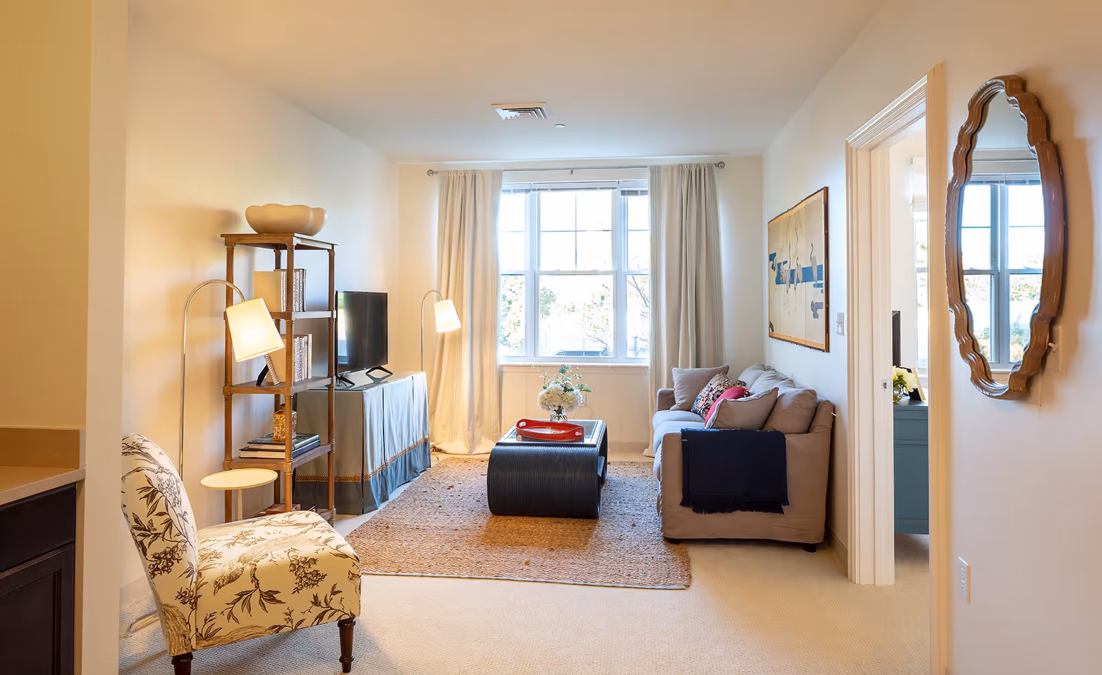 A cozy living room with a beige sofa adorned with cushions and a dark throw blanket, a black coffee table with a red tray and a flower arrangement, a patterned armchair, a wooden shelving unit with a TV, two floor lamps, a large window with cream curtains, a wall mirror, and a framed artwork on the wall.