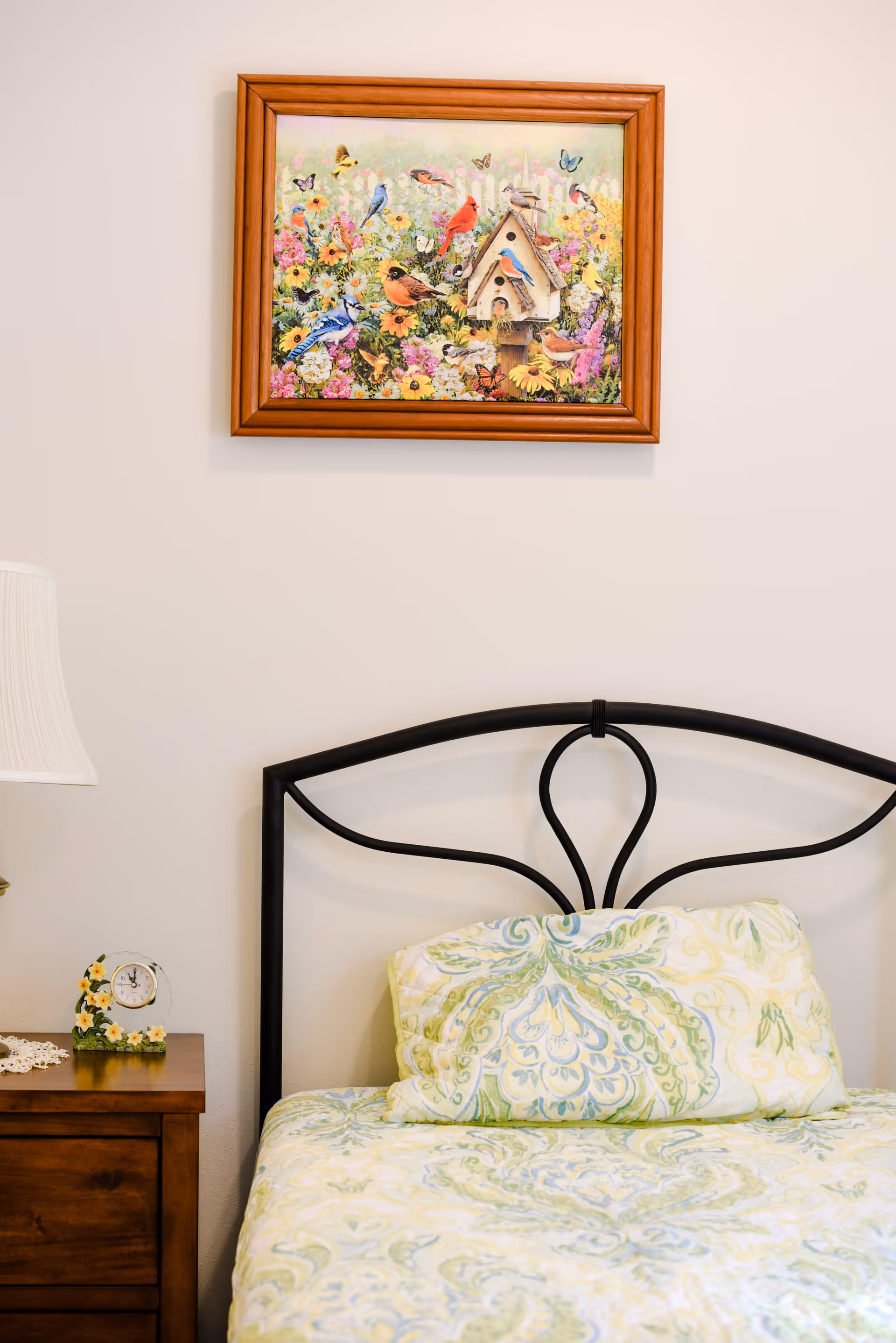 A bedroom scene with a bed featuring a black metal headboard and a pillow with a green and blue floral pattern. Above the bed is a framed picture of colorful birds and butterflies among flowers. To the left of the bed is a wooden nightstand with a small decorative clock and a white lampshade.