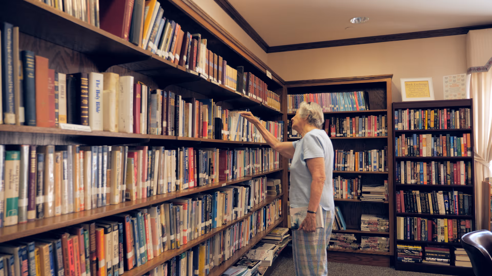 An elderly woman in a light blue shirt and plaid pants stands in a library room reaching for a book on a wooden bookshelf filled with various books. The room has multiple bookshelves, a window with curtains, and a framed sign on the wall.