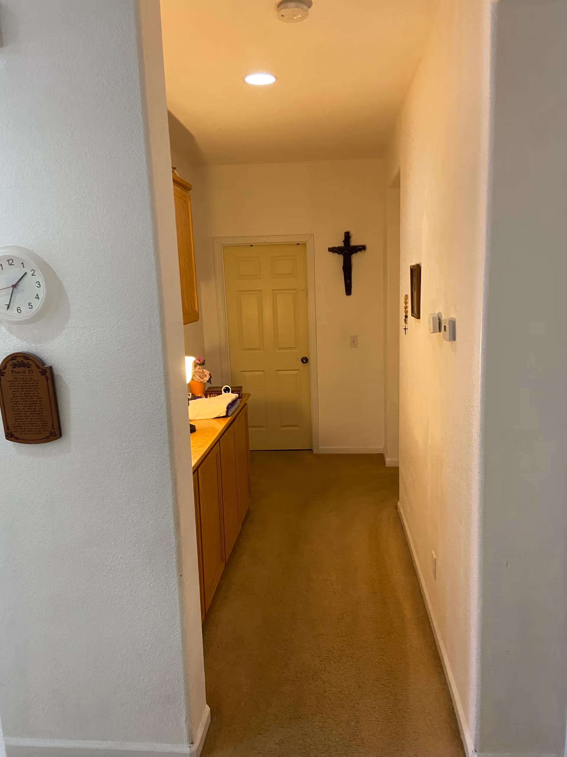 A narrow hallway with beige carpet and white walls. On the left side, there is a wooden cabinet with items on top, and a wall clock and a plaque are mounted on the wall. At the end of the hallway, there is a closed door and a crucifix hanging on the wall to the right of the door. The hallway is lit by a ceiling light.
