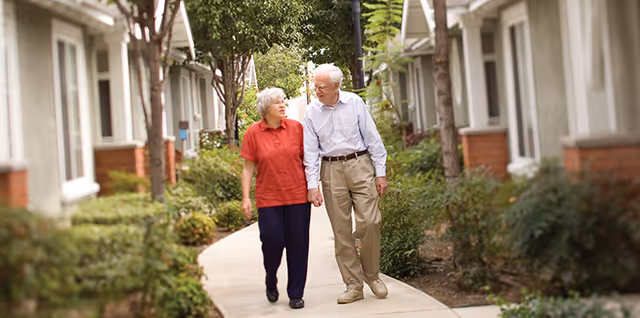 An elderly couple holding hands and walking along a paved pathway lined with greenery and trees, with residential buildings on either side.