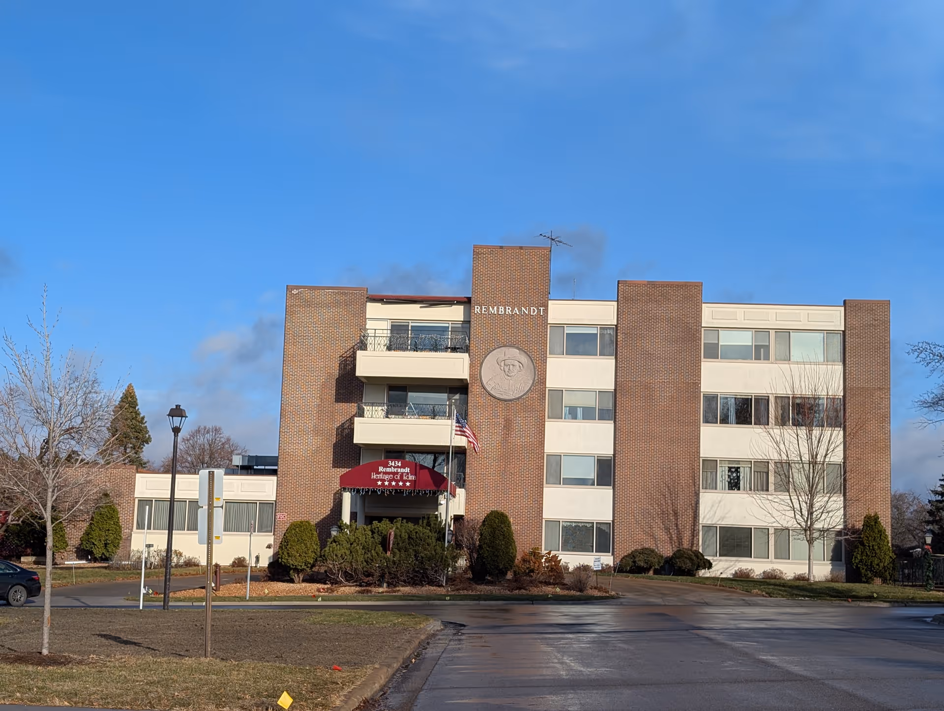 Front view of a four-story brick senior living building labeled 'Rembrandt' with balconies, an entrance awning, an American flag, and landscaped shrubs under a clear blue sky.